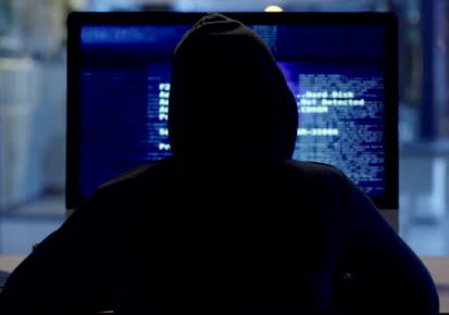 Back of hacker or cyber criminal stealing information online sitting at a computer at night. Hooded mysterious person coding spyware, malware and viruses to commit fraud, theft and phishing scam